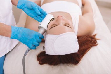 Young woman relaxing at beauty clinic while professional cosmetologist using  massage device on her face