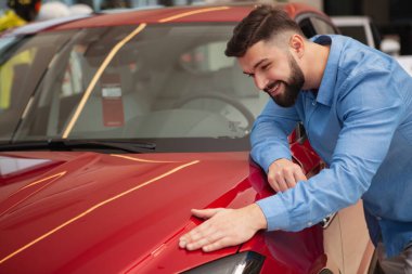 Mutlu yakışıklı sakallı adam gülümsüyor, araba galerisinde satışta olan güzel kırmızı otomobili inceliyor.