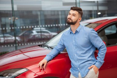 Genç yakışıklı sakallı adam otomobil galerisinde bir arabaya yaslanıyor, hayal kurarak yüzünü çeviriyor.