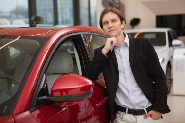 Çekici, genç, zarif bir adam. Hayallere dalmış, otomobil galerisinde yeni bir arabaya yaslanıyor.