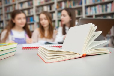 Seçici bir şekilde bir kitaba odaklan, arka planda birlikte okuyan üç genç kız öğrenci. Eğitim, arkadaşlık kavramı