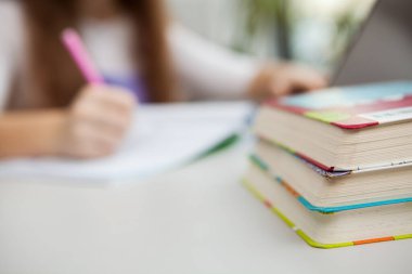 Kütüphanedeki ya da sınıftaki bir öğrenci masasının üzerine yığılmış bir yığın kitap. Tanımlanamayan bir kız arka planda bilgisayar üzerinde çalışıyor, kütüphanede okuyor.