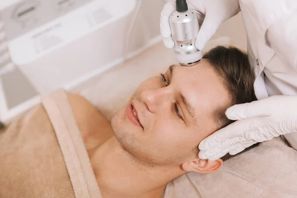 Professional cosmetologist using rf-lifting device on the face of handsome young male client