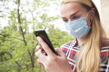 Dışarıda tıbbi maske takan genç bir kadın akıllı telefonunu kullanıyor. Sosyal medya, mobil uygulama kavramı