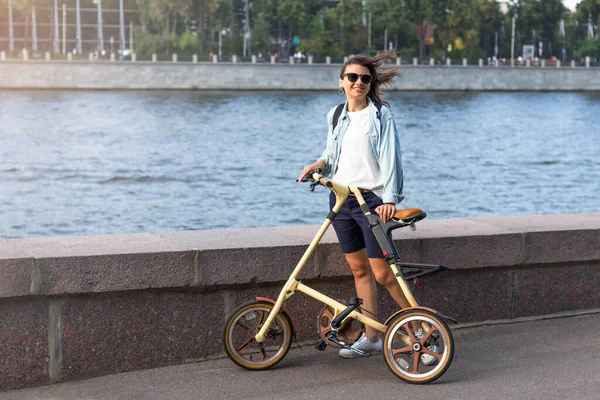 Güneş gözlüklü güzel beyaz kadın geniş bir nehrin kenarında duruyor. Konforlu sarı bir bisikletin ellerinde. Yaz ya da bahar yaşam tarzı.