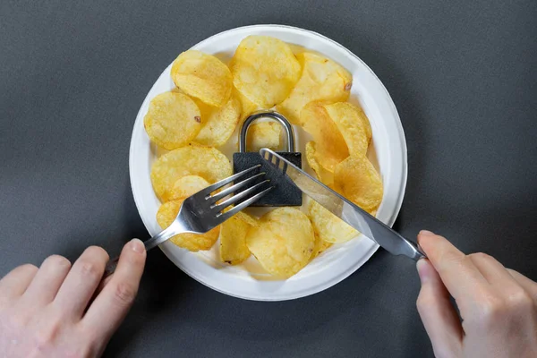 Çatal ve bıçaklı beyaz eller kilidi kesmiş. Gri arka planda beyaz tabakta kilitli lezzetli cipsler. Abur cubur.