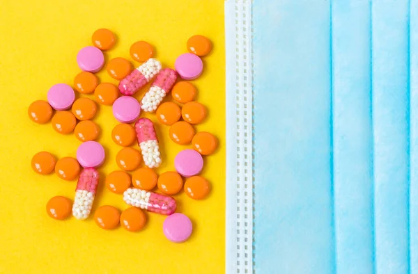 Çok renkli haplar ve parlak sarı arka planda tek kullanımlık mavi bir maske. Hastalık ilacı. Coronavirus virüsü hastalığından alınan ilaçlar ve sağlık için vitaminler. Kapatın. Boşluğu kopyala.