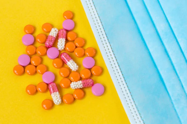Multi-colored pills and a blue disposable mask on a bright yellow background, top view. Medication for the disease. Drugs from coronavirus virus disease and vitamins for health. Close up. Copy space.