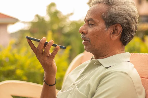 Senior man using virtual assistant in phone - Indian elder man using voice command on mobile - old man talking on smartphone at outdoor.