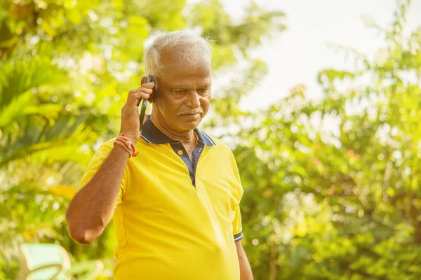 Elder man busy in mobile - Healthy senior Indian on phone outdoor at park, Morning - Happy male parent talking on phone.