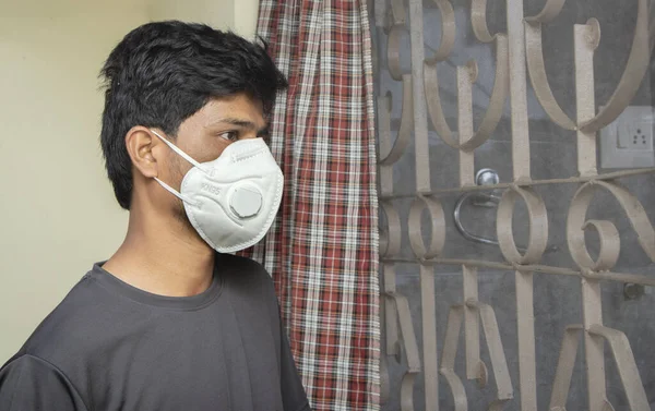 Молодой человек в медицинской маске смотрит через окно - концепция домашней изоляции или карантина в связи с ковидом 19 или вспышкой коронавируса — стоковое фото