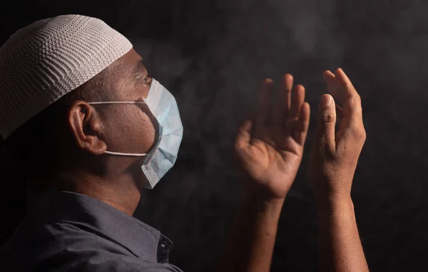 Hombre musulmán con máscara médica y gorra orando a Dios en cuarto oscuro para proteger o salvar de la crisis de covid-19 o coronavirus - espiritualidad y concepto de rendición . — Foto de Stock