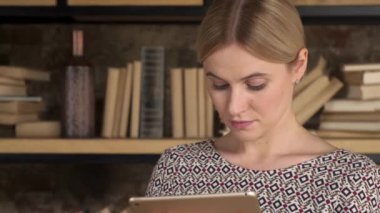 Kitaplığın yanında tablet kullanan güzel bir kadın. Güzel kız modern evdeki bilgisayar ekranına dokundu.