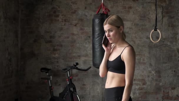 健身女人打电话给健身俱乐部的手机 年轻女子坐在体育馆里 在健身俱乐部用智能手机打电话的漂亮姑娘 — 图库视频影像