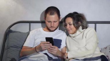 Mutlu genç bir çift cep telefonlarına bakarken evde yatakta uzanıyorlar.