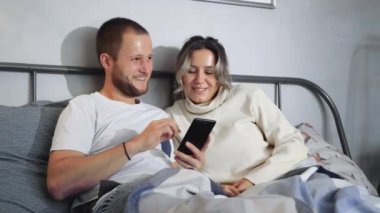 Mutlu Genç Çift Gece Yatağında Akıllı Telefon Kullan. Karı koca akıllı telefon kullanarak evde oturup gülüyorlar.