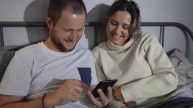 Mutlu Genç Çift Gece Yatağında Akıllı Telefon Kullanıyor, Ekranı Paylaşıyorlar, Sosyal ağlar Arasında Göz gezdiriyorlar, Resimleri Paylaşıyorlar, İnternet Kullanıyor ve Eğleniyorlar. Mavi Renkli Üst Görünüm Kamerası