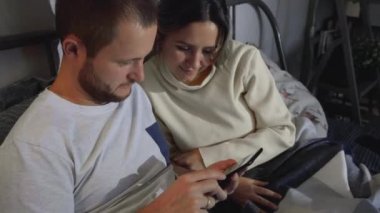 Akıllı telefondan ortak fotoğraflar izleyen bir çift. Mutlu çift fotoğraflara bakmak için akıllı telefon kullanıyor. Yatakta yatıyorlar. Genç adam ve kadın konuşuyor, birlikte vakit geçiriyorlar..