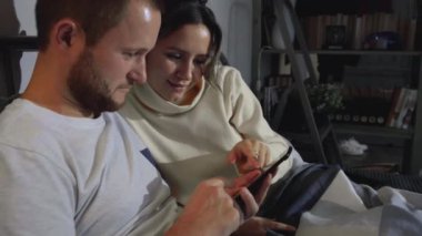 Mutlu çift fotoğraflara bakmak için akıllı telefon kullanıyor. Yatakta yatıyorlar. Mutlu genç bir çift cep telefonlarına bakarken evde yatakta uzanıyorlar.
