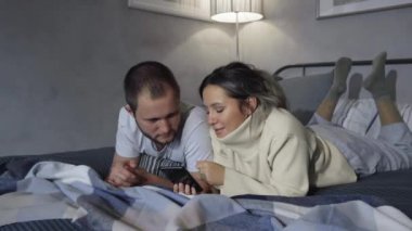 Mutlu Genç Çift Gece Yatağında Akıllı Telefon Kullanıyor, Ekranı Paylaşıyorlar, Sosyal Ağ Taraması, Fotoğrafları Paylaşıyorlar, İnterneti Kullanıyorlar ve Eğleniyorlar. 