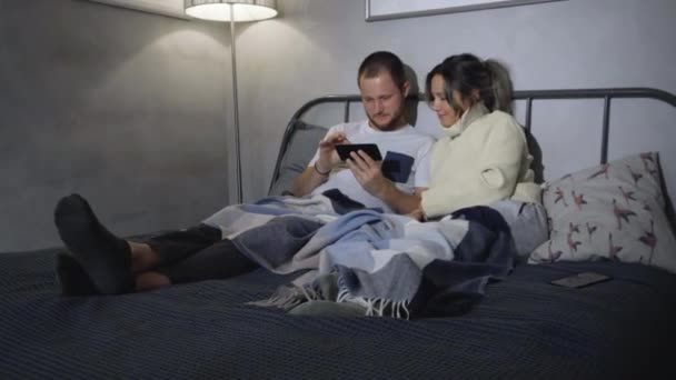 夫婦が携帯電話を手にベッドに横たわっている ベッドルームでポータブルデバイスを使用して若いカップル — ストック動画