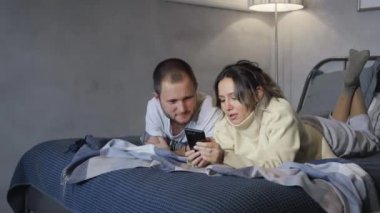 Ev 4K 'deki yatakta cep telefonu kullanan çift. Kafası karışmış genç bir çift evde yatakta oturan bir cep telefonunu işaret ederken tartışıyor.