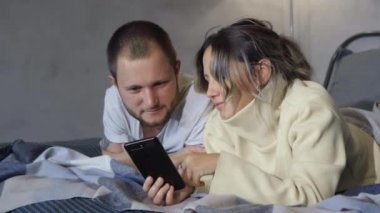 Evli çift akıllı telefonlar kullanarak yatakta yatıyor. Bir çift eş yatakta uzanıp akıllı telefonları izliyor. Sosyal ağlarda iletişim. Kadın ve erkek evde yatıyor.