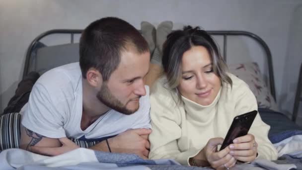 笑着的年轻夫妇深夜坐在床上 用智能手机拍片 — 图库视频影像