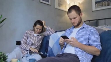 Genç ve mutlu bir çift ellerinde telefonla ekranlara bakıyor ve modern oda dairesinde oturuyorlar. Kadın ve erkek evde.