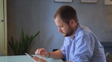 Düşünceli genç iş adamı sandalyede oturuyor ve büyük bir pencereye karşı ofiste tablet kullanıyor. Ağır çekimde çekim takip ediliyor.