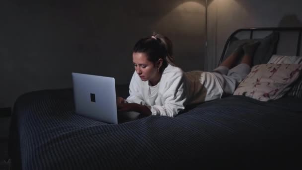 女性はノートパソコンを持ってベッドの上にいます 女の子学生は インターネットの先生とオンラインヘッドフォンの研究を着用言語の話は ラップトップを見て学ぶ 焦点を当てた若い女性はビデオ通話を行う — ストック動画