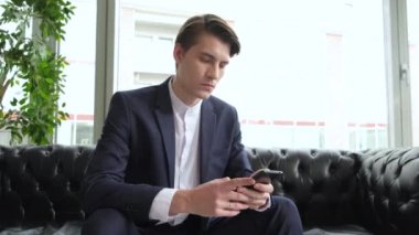 Erkek milenyum profesyoneli ofiste modern akıllı telefon mesajı tutuyor. Genç işadamı iş zamanı yönetim organizasyonu konsepti için yardımcı mobil uygulamalar kullanıyor. Çalışma masasında oturuyor..