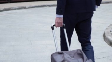 Güneşli bir günde şehir caddesinde yürüyen ve bavulu tekerlekli sandalyeyle çeken başarılı iş adamının ayakları. Kendine güvenen adam uçağına doğru gidiyor. Kentsel çevreye ayak basan ayakkabılı erkek bacakları. Yavaş çekim