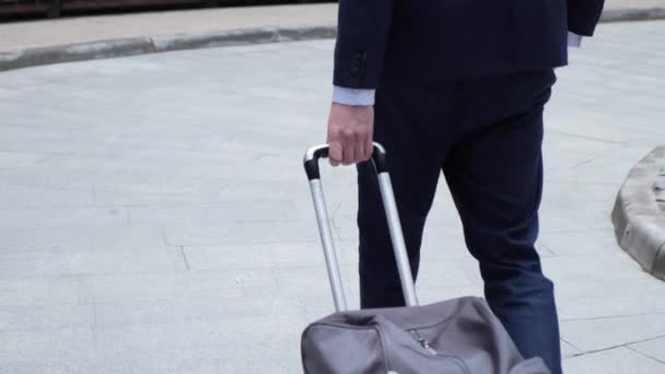 成功したビジネスマンの足は街の通りを歩くと晴れた日に車輪の上にスーツケースを引っ張っ 飛行中の男を自信を持って 都市環境に足を踏み入れる靴の男性の足 スローモ — ストック動画