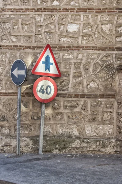 three traffic signs next to a stone wall of a church on a street.