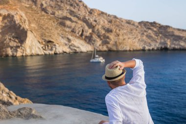 Şapkalı erkek arka siluet kayalık sahil ve bir yat ile mavi deniz manzarasına sahiptir