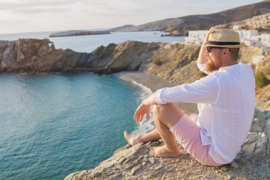 Bir sakallı hippi erkek tatil dik bir sahil üzerinde oturur ve mesafe içine görünüyor