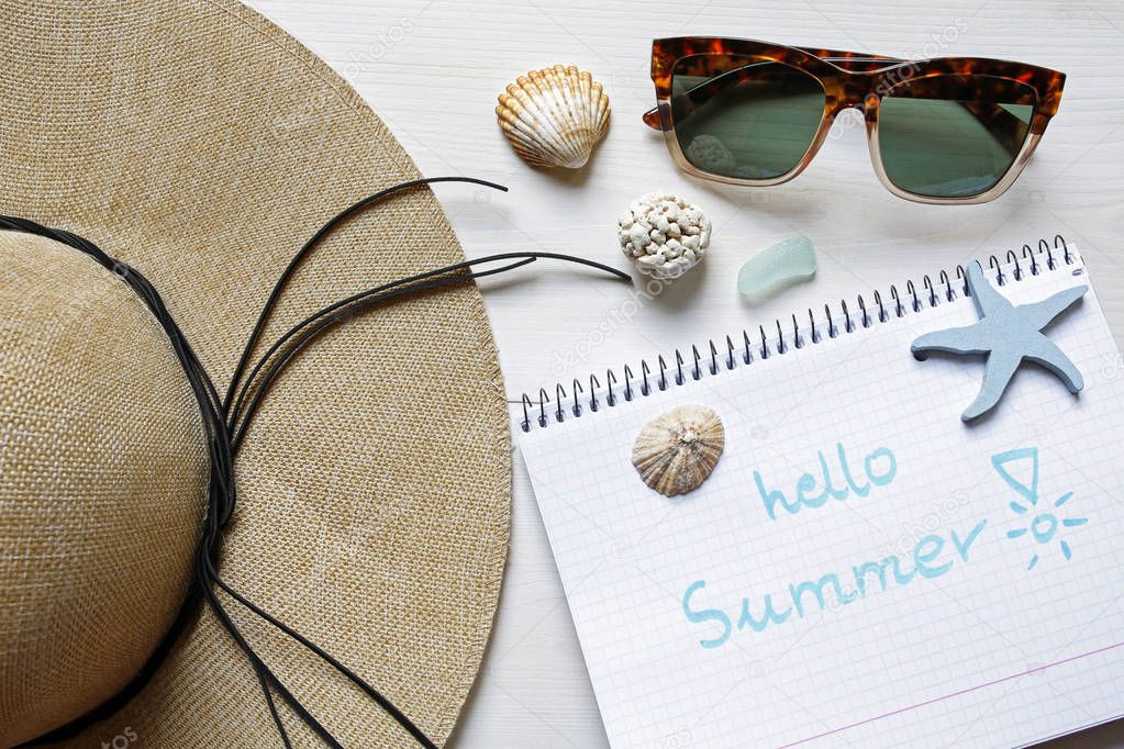 hello summer concept- straw hat, seashells, sunglasses and notebook on white wooden background