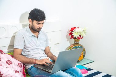Genç Hintli adamın laptopuyla meşgul olduğu yatak odasında dinlenirken ofis işleriyle meşgul olduğu, evden çalışan serbest çalışan. Bilgisayarda daktilo eden genç bir erkek öğrenci günlük giysiler giyiyor..