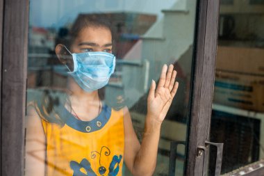 Coronavirus. Corona virüsünün hasta genç kızı pencereden dışarı bakıyor ve maske takıyor ve evdeki hastalıktan korunuyor. Karantinaya alın. Hasta enfeksiyonu önlemek için izole edilmiş..