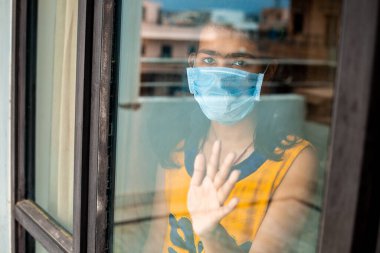 Coronavirus. Corona virüsünün hasta genç kızı pencereden dışarı bakıyor ve maske takıyor ve evdeki hastalıktan korunuyor. Karantinaya alın. Hasta enfeksiyonu önlemek için evde izole edilmiş. Coronavirus. Corona virüsünün hasta genç kızı