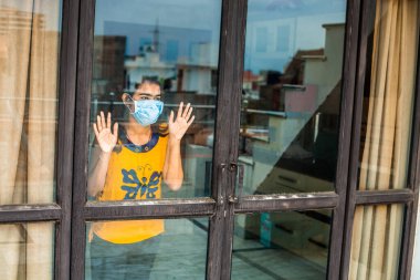 Coronavirus. Corona virüsünün hasta genç kızı pencereden dışarı bakıyor ve maske takıyor ve evdeki hastalıktan korunuyor. Karantinaya alın. Hasta enfeksiyonu önlemek için izole edilmiş..