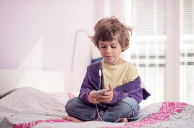 Küçük çocuk yatak dinleme müzik onun akıllı telefon üzerinde oturan.