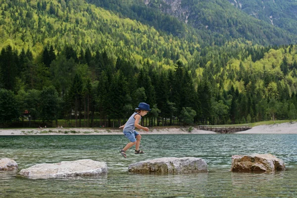 Adorable young boy with a hat crossing river or water jumping from rock to rock. Crossing the gap, freedom, liberation, success, avoiding danger, courage concept