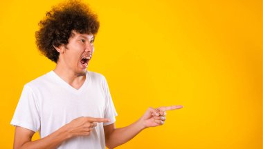 Asya portresi mutlu kıvırcık saçlı genç adam beyaz tişört parmakları gösteren kopya alanı izole edilmiş sarı arkaplan