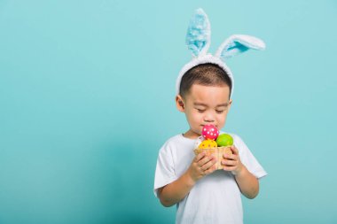 Asyalı şirin çocuk gülümsüyor tavşan kulağı ve beyaz tişört giyiyor. Mavi arka planda paskalya yumurtası dolu bir sepeti tutmak için bekliyor.