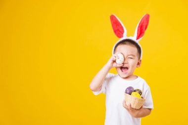 Paskalya bayramında beyaz tişört ve tavşan kulağı giyerek mutlu mesut gülümseyen küçük Asyalı çocuk paskalya yumurtaları taşıyor, sarı arka planda fotokopi çekimleri yapıyor.