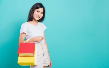 Portre mutlu Asyalı genç bir kadın beyaz tişört giyiyor, elinde alışveriş çantaları var, çok renkli ve kameraya bakıyor, mavi arka planda bir stüdyoda fotoğraf çekiyor.