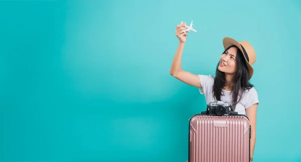 Traveler tourist happy Asian beautiful young woman, holidays travel concept, her holding fly toy aircraft her have suitcase bag and photo mirrorless camera, shoot photo in studio on blue background