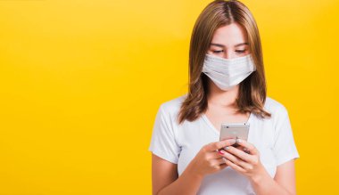 Asya Tayland portresi maske takan güzel, mutlu genç bir kadın, akıllı telefon sağlık hizmetleri ile ilgili haberleri okuyor, sarı arka planda izole edilmiş bir stüdyo çekimi, fotokopi alanı ile